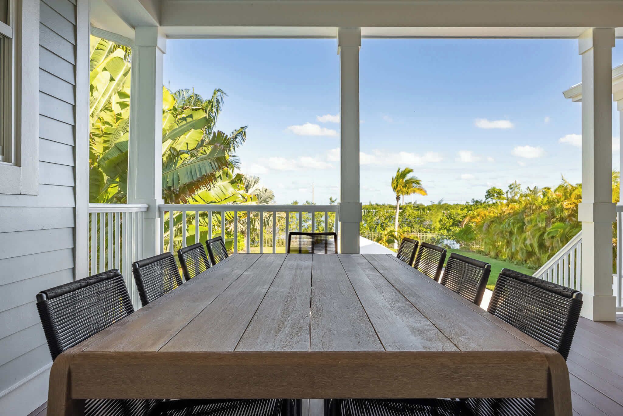 Une table en bois, entourée de six chaises, est placée sur une véranda d’où l’on peut admirer un jardin luxuriant et des palmiers.