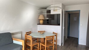 Un espace de vie compact comprenant une table de salle à manger ronde, des chaises en bois, un canapé gris, un coin cuisine avec une lampe suspendue et une porte donnant sur une autre pièce.