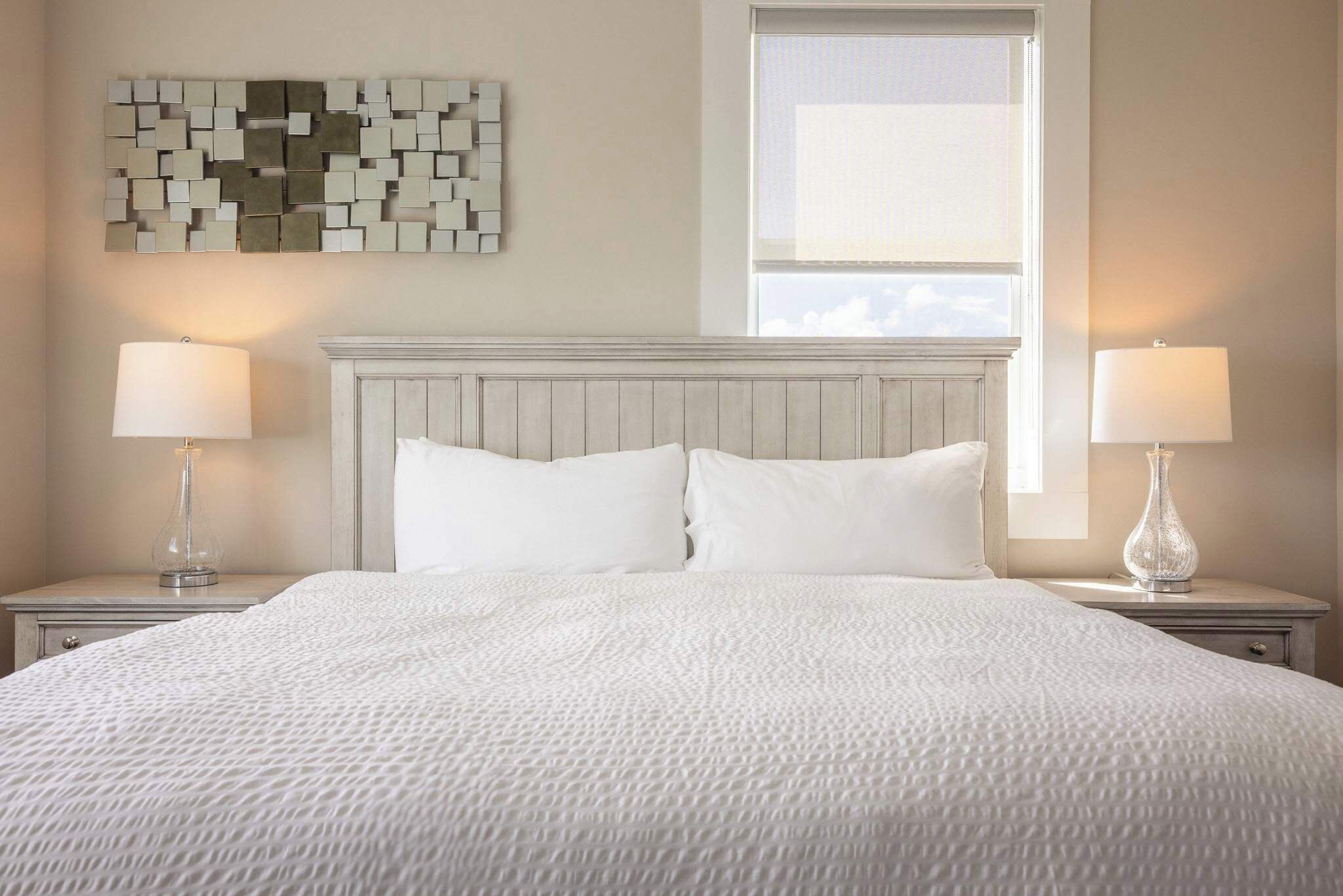 Una cama bien tendida con sábanas blancas, dos lámparas de noche, un cabecero de madera y una obra de arte en la pared.