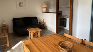A modern living room with a black sofa, wooden coffee table, and a dining table. There is a kitchen visible through an open doorway.