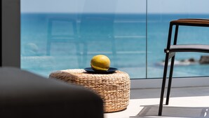 Un otoman de mimbre con una fruta amarilla encima, una silla negra y una vista al océano a través de una ventana de vidrio.