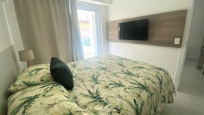 A hotel room with a bed, a TV mounted on the wall, a window with curtains, and a nightstand with a lamp. - In Mare praia de Cotovelo por Carpediem (Parnamirim)