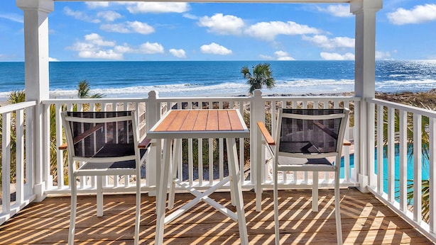 A covered outdoor seating area with a wooden table and two chairs, overlooking a beach with the ocean and a clear sky. - "Mermaid's Delight" Oceanfront Beach House w/pool (Murrells Inlet)