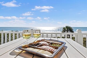 A tray of food and two glasses of white wine on a wooden table with a view of the ocean. - Beachfront House - Dogs OK - Pool, Near Golf (Murrells Inlet)