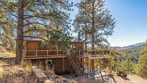 A wooden cabin with a deck and staircase, surrounded by trees and hills.