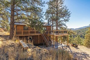 Una cabaña de madera con terraza y escalera, rodeada de árboles y colinas.