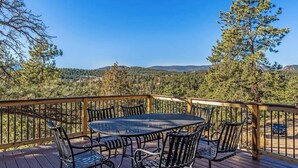 A wooden deck with a round table and chairs, surrounded by a metal railing, overlooking a forested landscape.