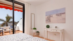 A bedroom with a bed, a desk, a chair, a potted plant, a mirror, and a beach view painting. - "Piece of Ocean by Mea Substantia" (Playa de la Américas)