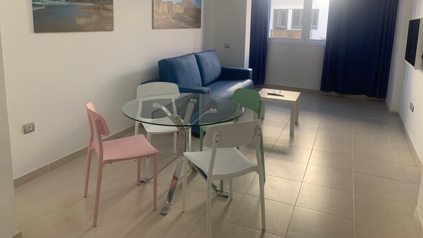 Ein modernes Wohnzimmer mit einem Glastisch, einem blauen Sofa und zwei Sesseln. An den Wänden hängen zwei Bilder und ein Fenster mit Vorhängen.