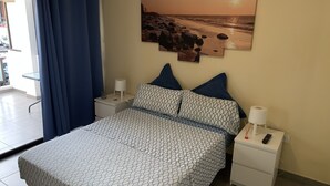 Ein Schlafzimmer mit einem Bett, zwei Nachttischen, einem Fenster mit Vorhängen und einem Wandbild mit einer Strandansicht.