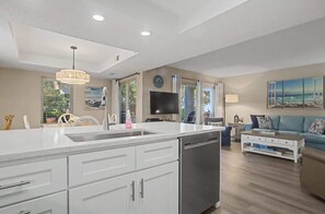 A modern kitchen with a large island, a dining area with a blue sofa, and a living room with a TV and artwork. - Luxury Villa, w/ Patio Steps to Beach Hilton Head (Hilton Head Island)