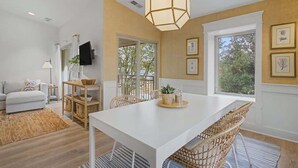 A dining area with a white table, wicker chairs, a hanging light fixture, and a view through large windows.