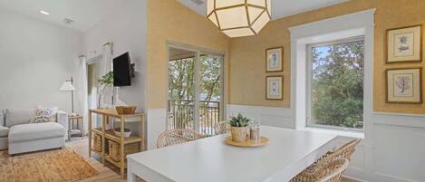 A dining area with a white table, wicker chairs, a hanging light fixture, and a view through large windows.