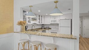 A kitchen with white cabinets, a granite countertop, a refrigerator, and two bar stools.
