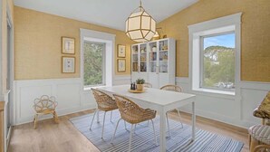 A dining area with a white table, wicker chairs, a hanging light fixture, and a cabinet with glass doors.