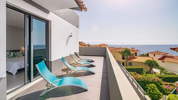 A balcony with a bed and lounge chairs, overlooking a coastal area with houses and the sea.
