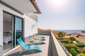 A balcony with a bed and lounge chairs, overlooking a coastal area with houses and the sea.