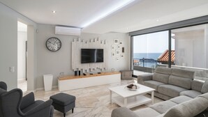 A modern living room with a large TV, grey sofas, a white coffee table, and a view of the sea.