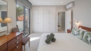 A modern bedroom with a large bed, a wooden dresser, a desk with a lamp, a sliding glass door to a balcony, and a wardrobe with a mirror.