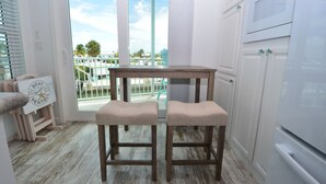A kitchen with a wooden table and two stools, a refrigerator, and a view of a pool and palm trees through a glass door. - 44ft Seawall at Sportsman's Paradise (Summerland Key)