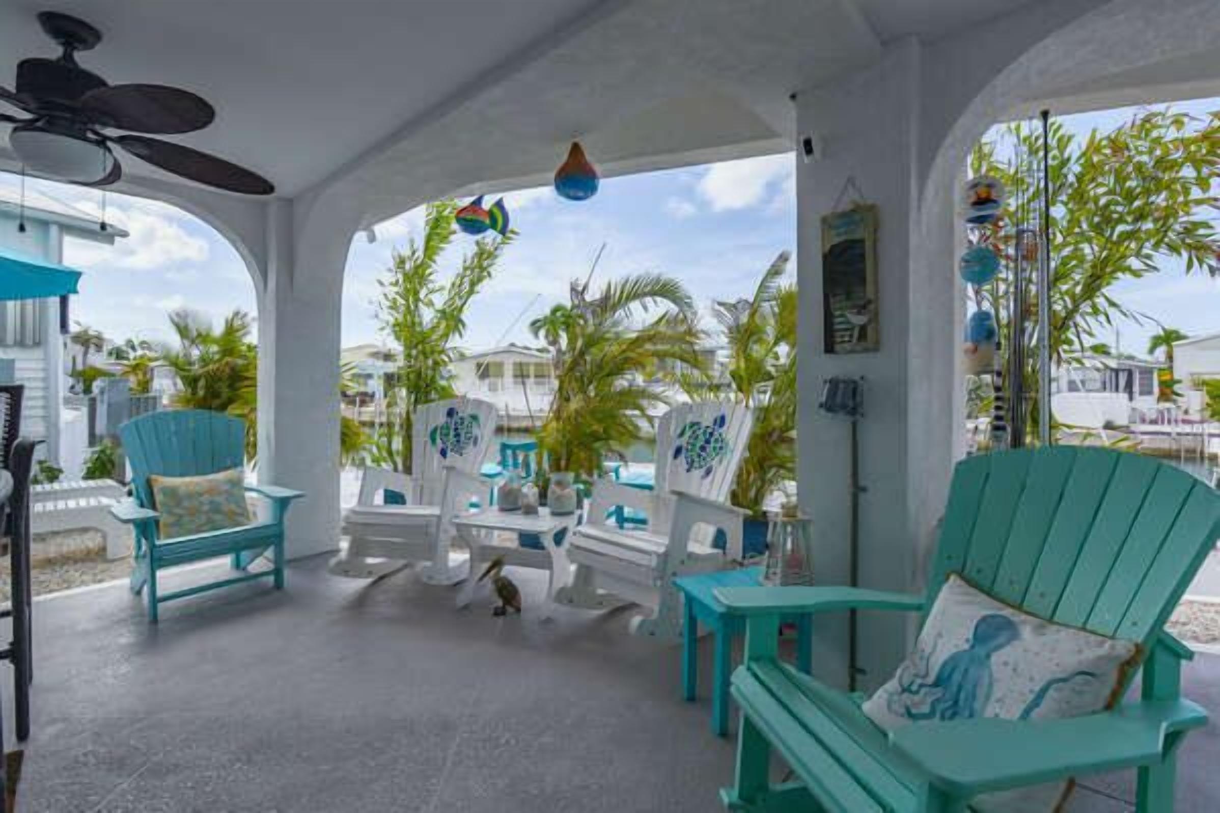 A covered outdoor seating area with turquoise chairs and tables, a ceiling fan, and a view of palm trees and buildings.
