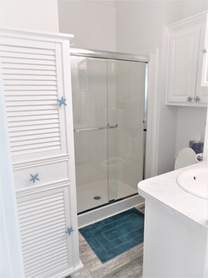 Ein Badezimmer mit Duschkabine aus Glas, weißen Schränken mit Seestern-Dekoration, einem weißen Waschbecken und einem blauen Teppich.