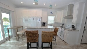 A modern kitchen with white cabinetry, a central island with bar stools, a built-in oven, and a large window with blinds. - 44ft Seawall at Sportsman's Paradise (Summerland Key)