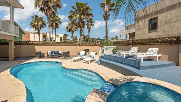 A swimming pool with lounge chairs and a deck area, surrounded by palm trees and a building with a balcony.
