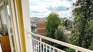 Un balcón con césped artificial, barandilla blanca y vista a una calle con coches aparcados y edificios.