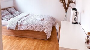 Un dormitorio con piso de madera, una cama con ropa de cama estampada, un escritorio con lámpara y una ventana con cortinas.