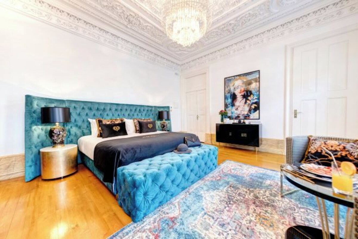 Un dormitorio con cabecera capitonada, una cama grande, una mesita de noche con lámpara, un aparador y un cuadro en la pared.