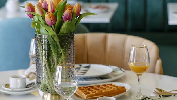 A dining table with a vase of tulips, a waffle, and a glass of orange juice. - 1877 Estrela Palace Hotel (Aveiro)