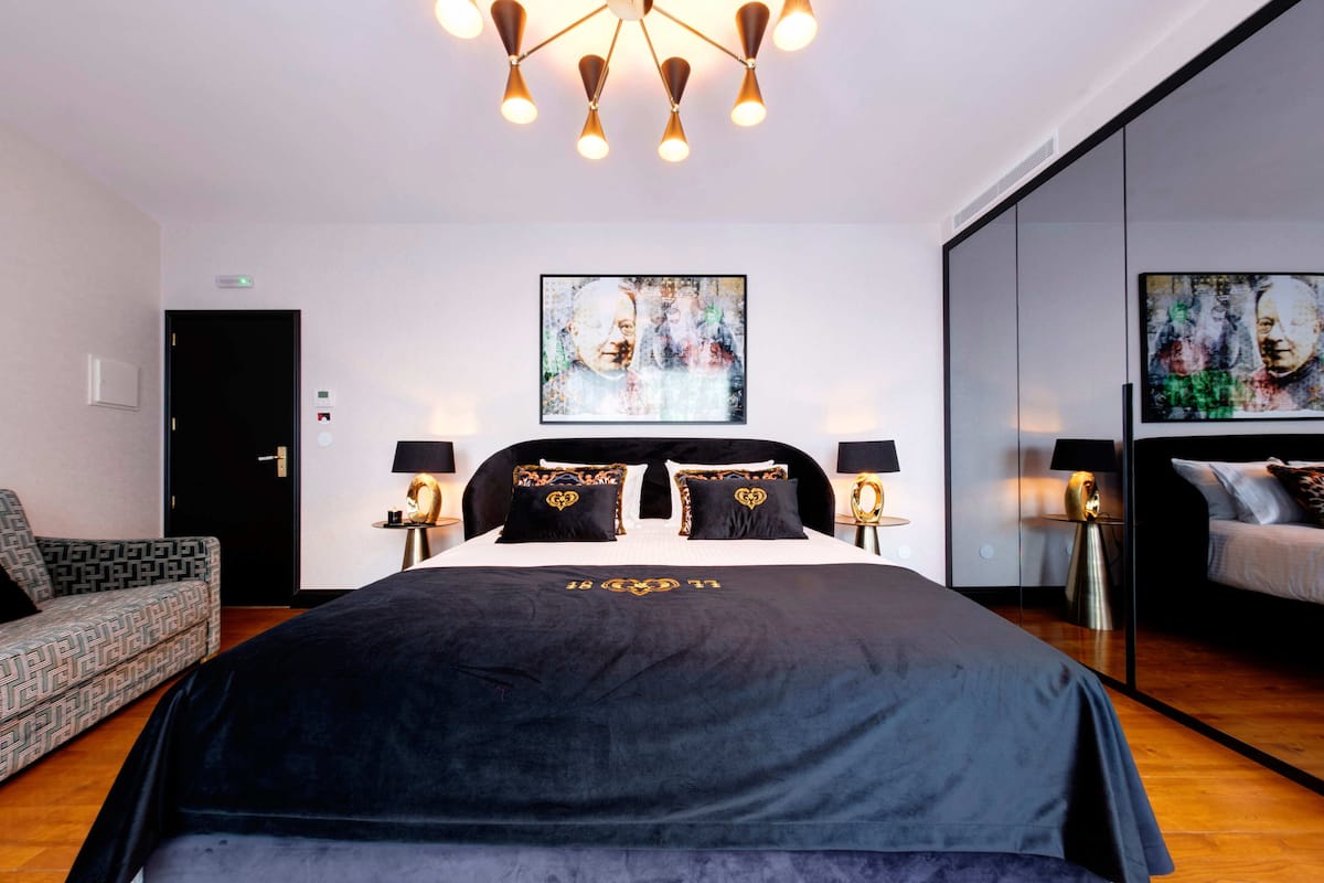 Una habitación de hotel moderna con una cama grande, dos mesitas de noche con lámparas, un sillón estampado y arte abstracto en las paredes.