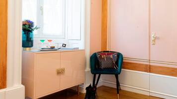 Quarto com uma grande janela, parede rosa, piso de madeira, cadeira azul-esverdeada e uma pequena mesa com um vaso e livros.