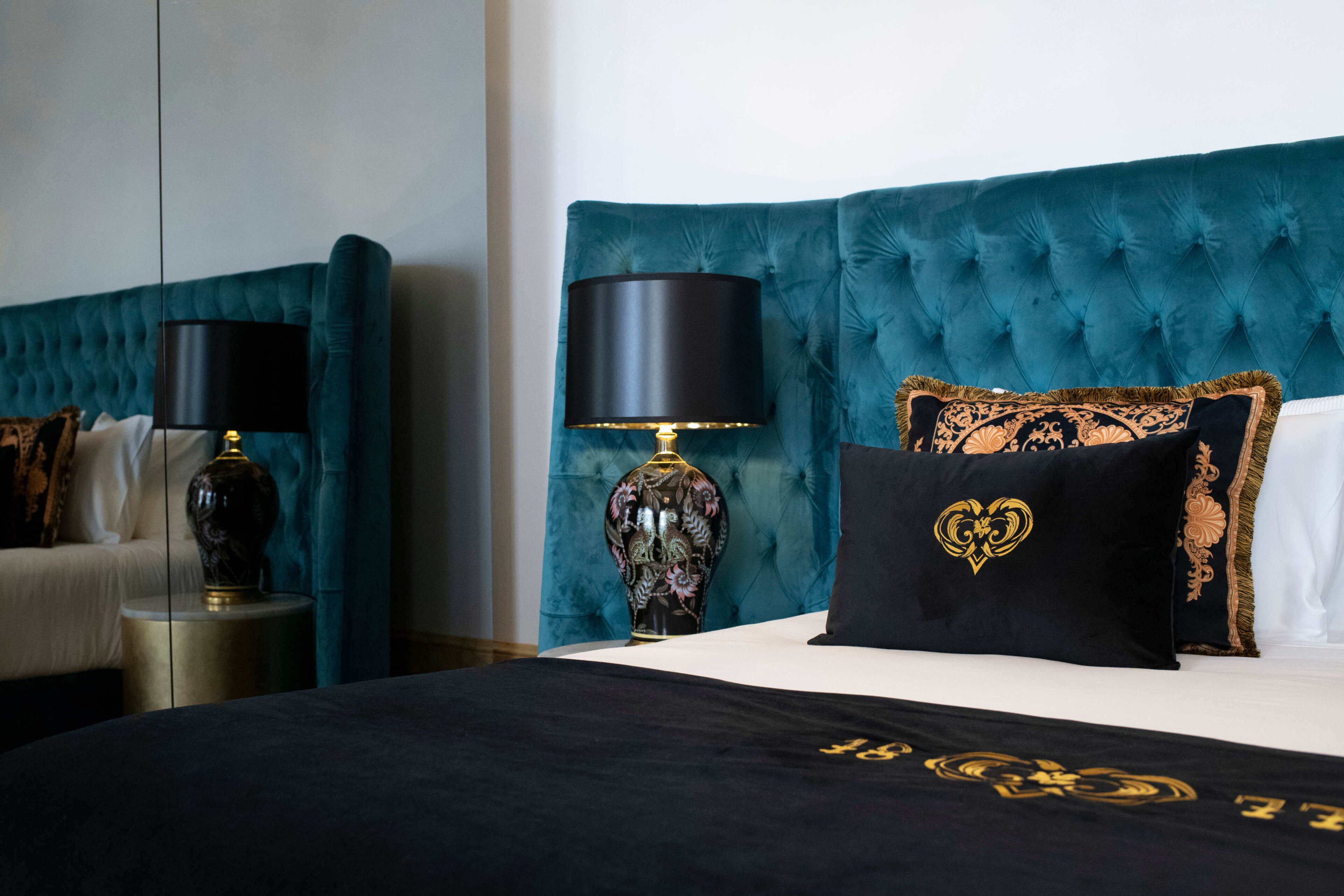 A hotel room with a tufted teal headboard, a black and gold decorative vase, and a black bedspread with gold embroidery.