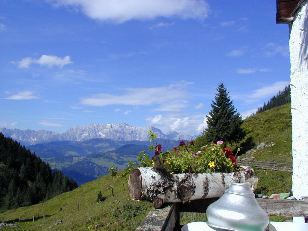 Vista de montanha com um vaso de madeira cheio de flores, um vaso de metal e um edifício branco com cerca de madeira.