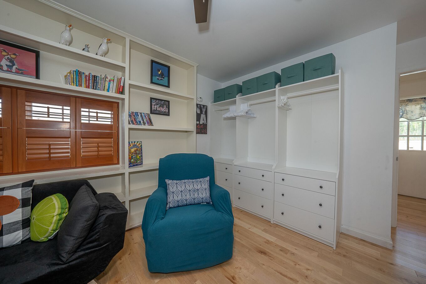 A living room with a teal armchair, a black sofa with patterned cushions, a white shelving unit with books and decorative items, and a white wardrobe with green storage boxes.