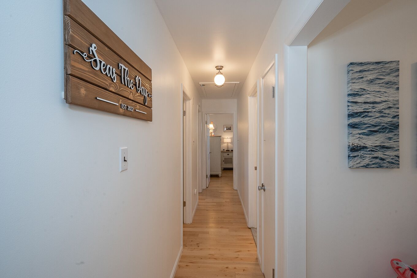 A hallway with wooden flooring, white walls, a ceiling light, and a sign on the left.