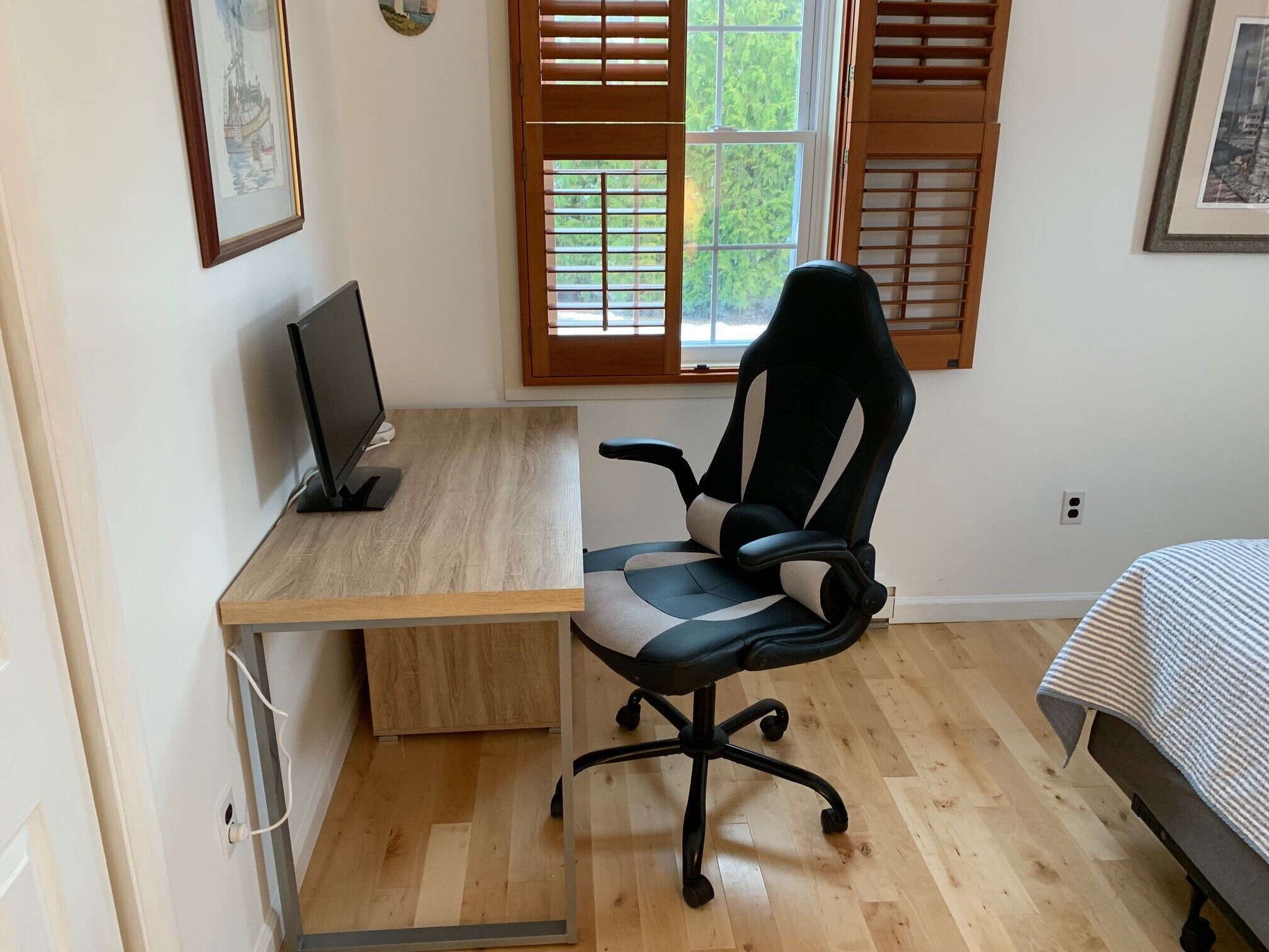 A bedroom with a wooden desk, a black office chair, a bed with a striped blanket, and a window with wooden shutters.