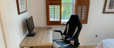A bedroom with a wooden desk, a black office chair, a bed with a striped blanket, and a window with wooden shutters.