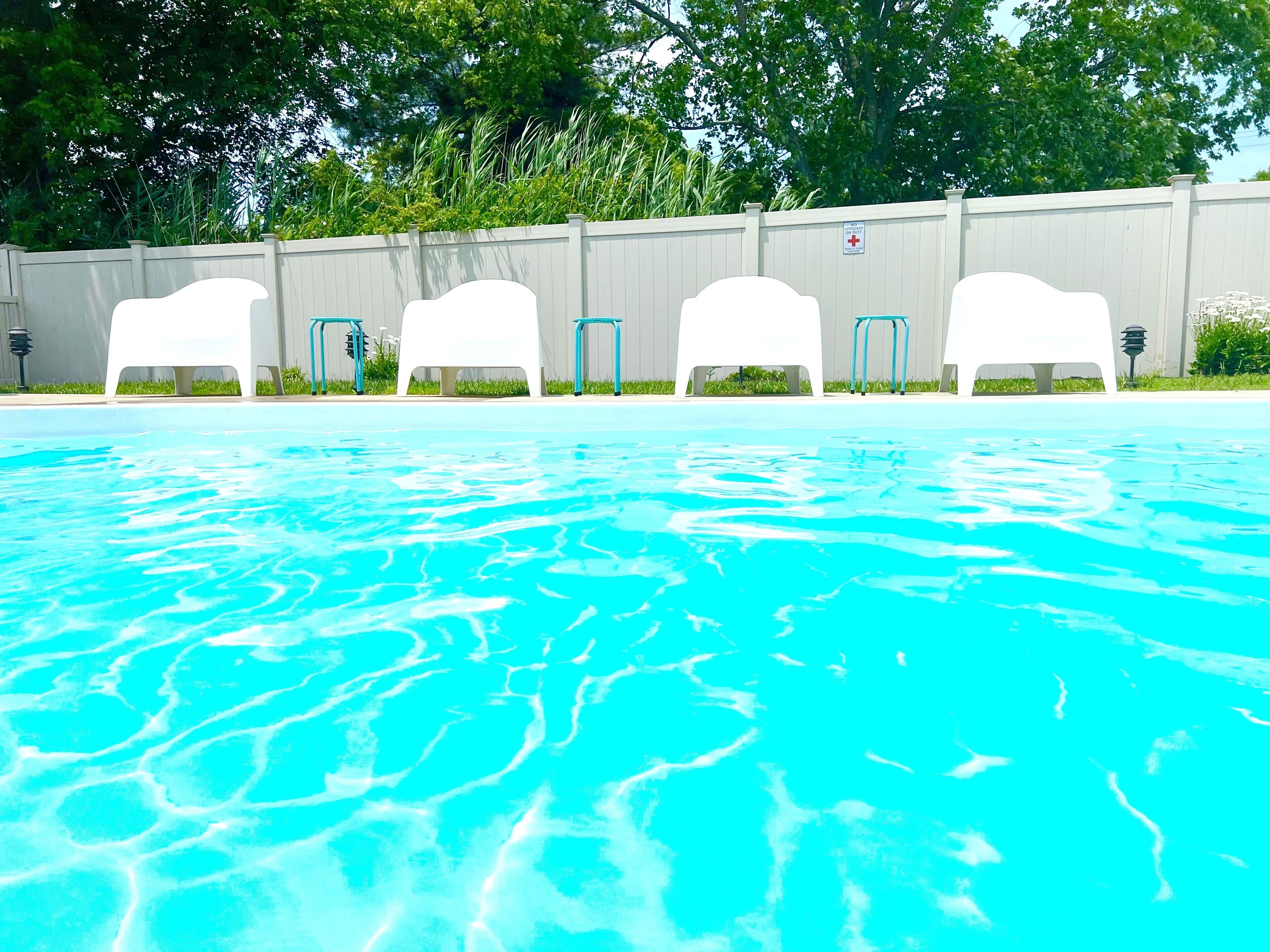 A swimming pool with clear blue water, surrounded by white lounge chairs and a white fence with greenery in the background.