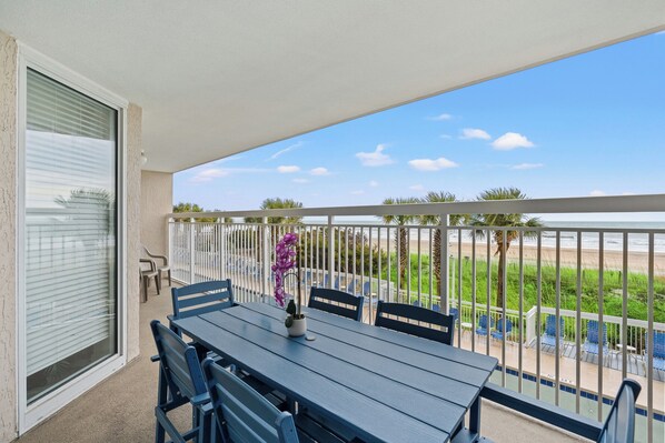A balcony with a table and chairs, a view of the beach, and a palm tree.