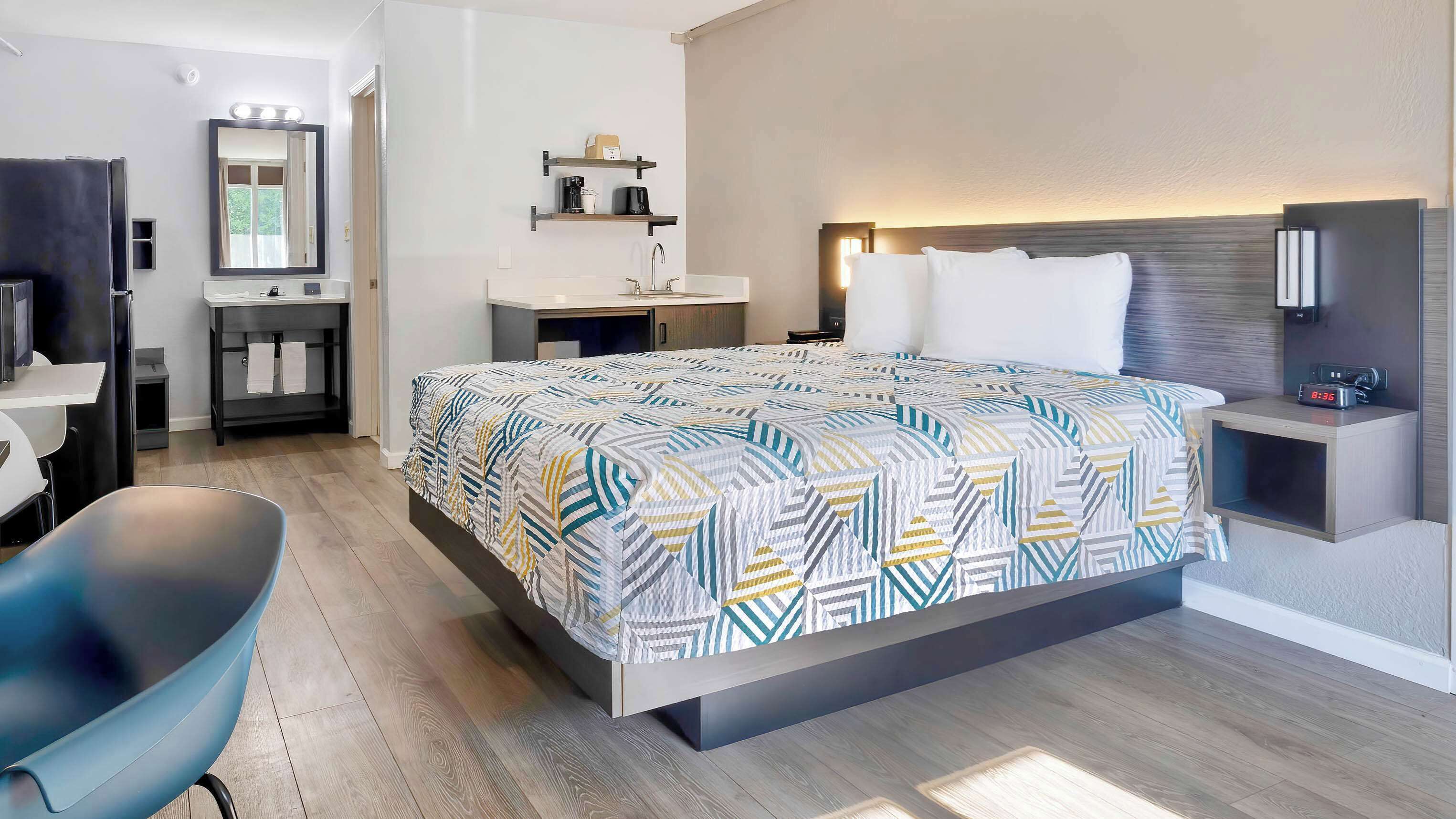 A modern hotel room with a large bed, a desk, a chair, a vanity with a mirror, and a bedside table with a lamp.