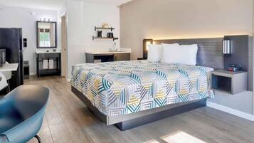 A modern hotel room with a large bed, a desk, a chair, a vanity with a mirror, and a bedside table with a lamp.