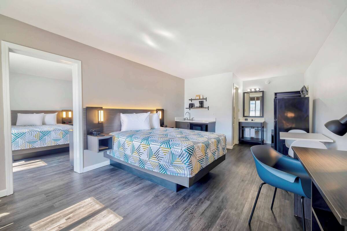A modern hotel room with a large bed, a desk, a chair, a mirror, and a small kitchenette.