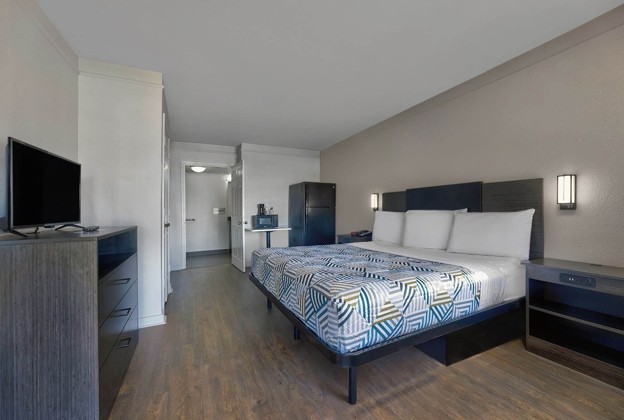 Quarto de hotel moderno com cama, mesa de cabeceira, televisão e uma pequena cozinha.