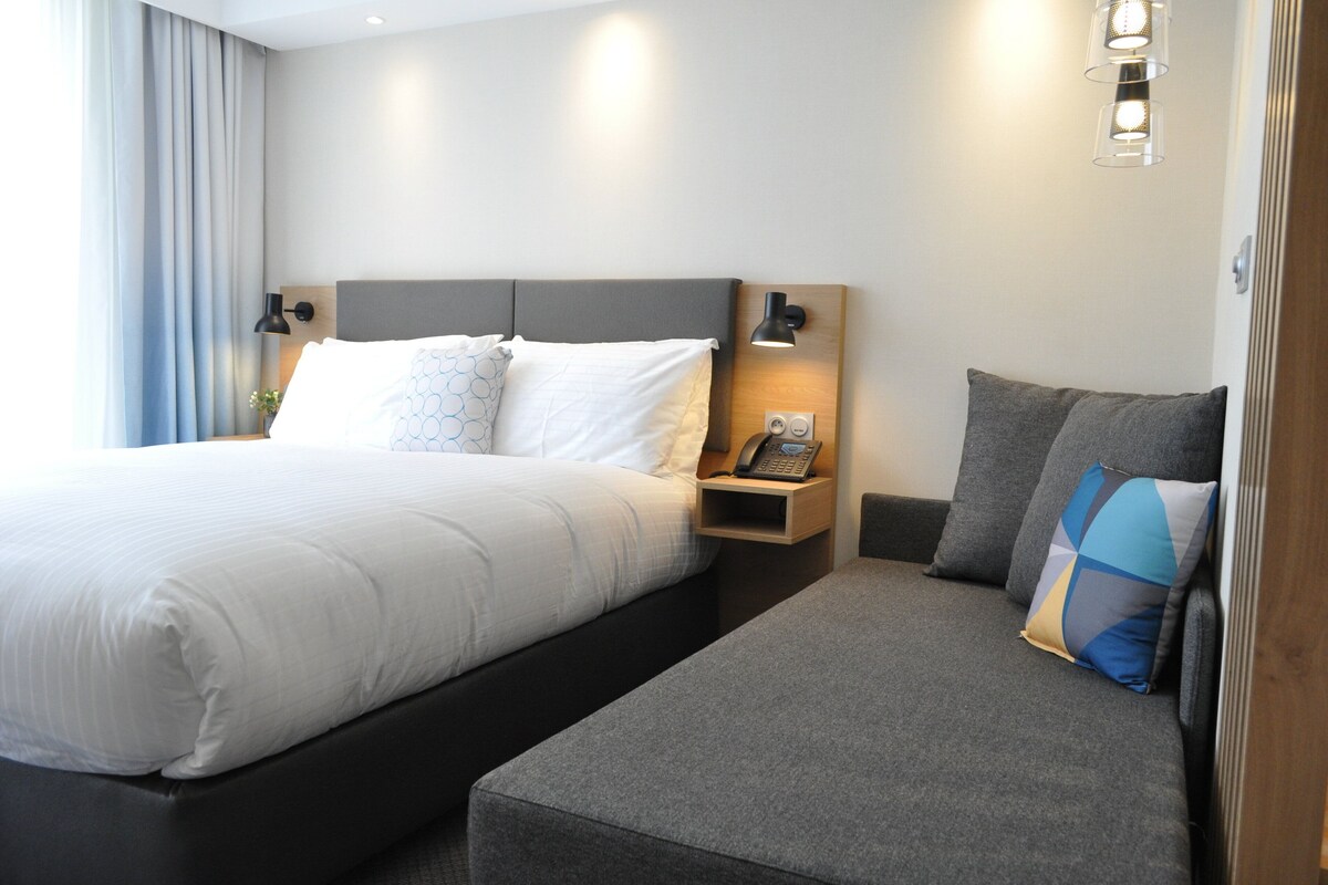 Ein modernes Hotelzimmer mit einem großen Bett, einem Nachttisch mit Lampe und einem kleinen Sofa mit Kissen.