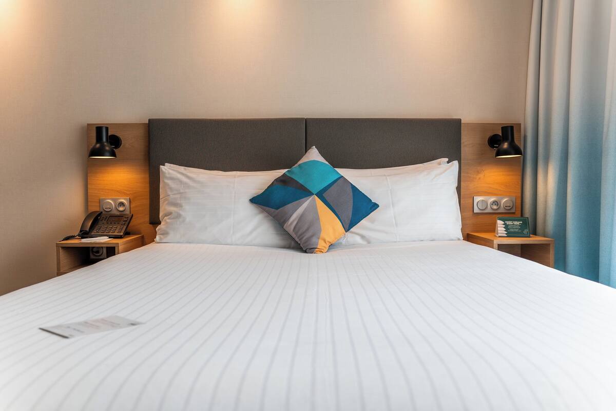 A neatly made bed with a white bedspread and two pillows, one with a geometric pattern. There are two bedside tables with lamps and a telephone on one.
