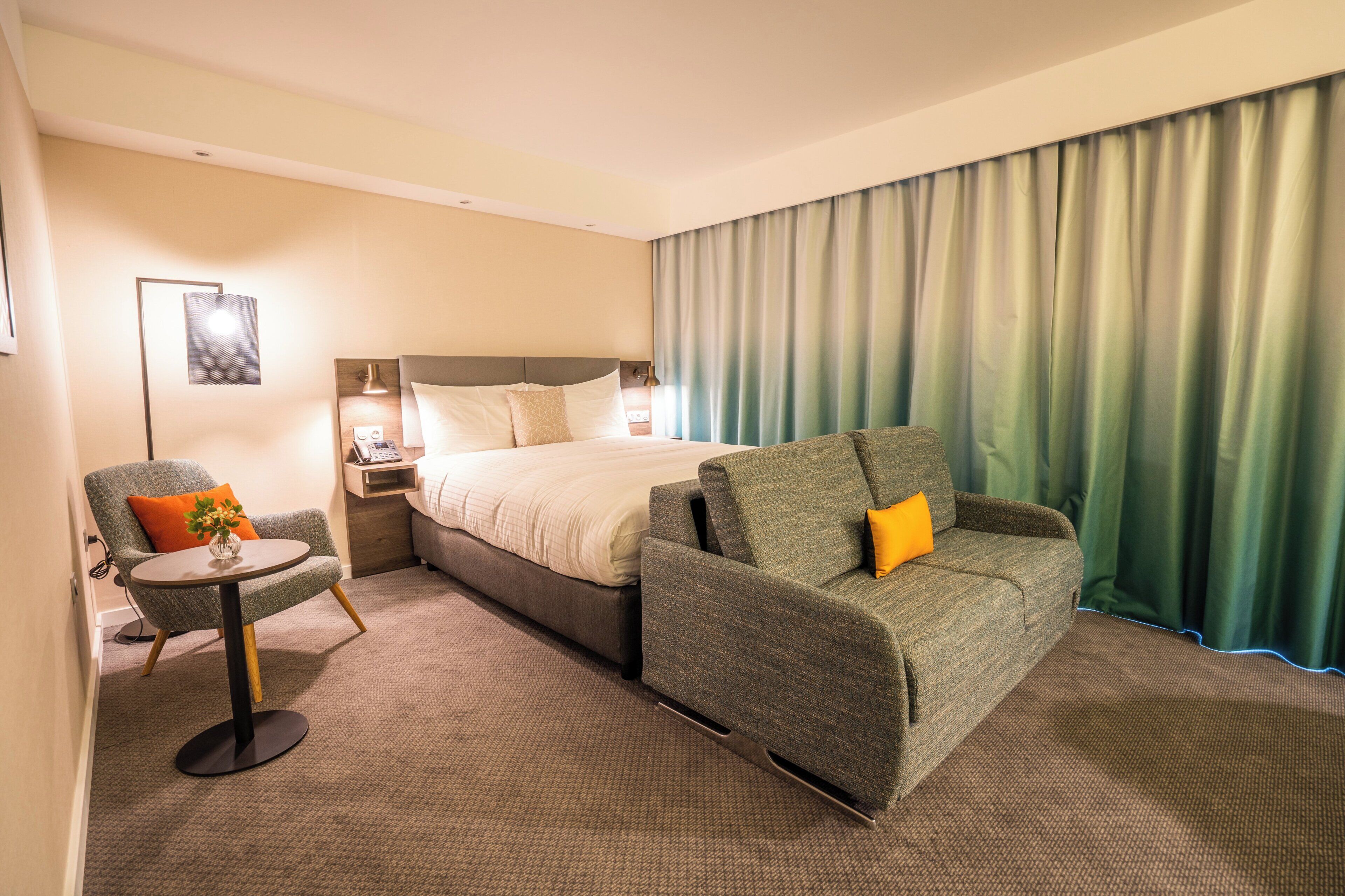 A hotel room with a bed, two armchairs, a small table, and a large window with curtains.