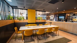 A modern conference room with a long table and yellow chairs, a bar area with a TV, and large windows with plants. - Holiday Inn Bordeaux-Merignac by IHG (Merignac)